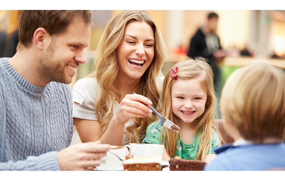 Glad familie med mor, far, datter og sønn som sitter å spiser kake og drikker kaffe på en kafe