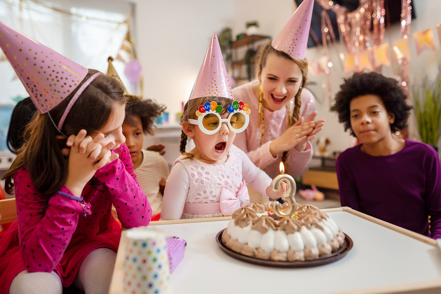 Fire barn i fargerike festhatter samles rundt en bursdagskake med et lys formet som nummer tre